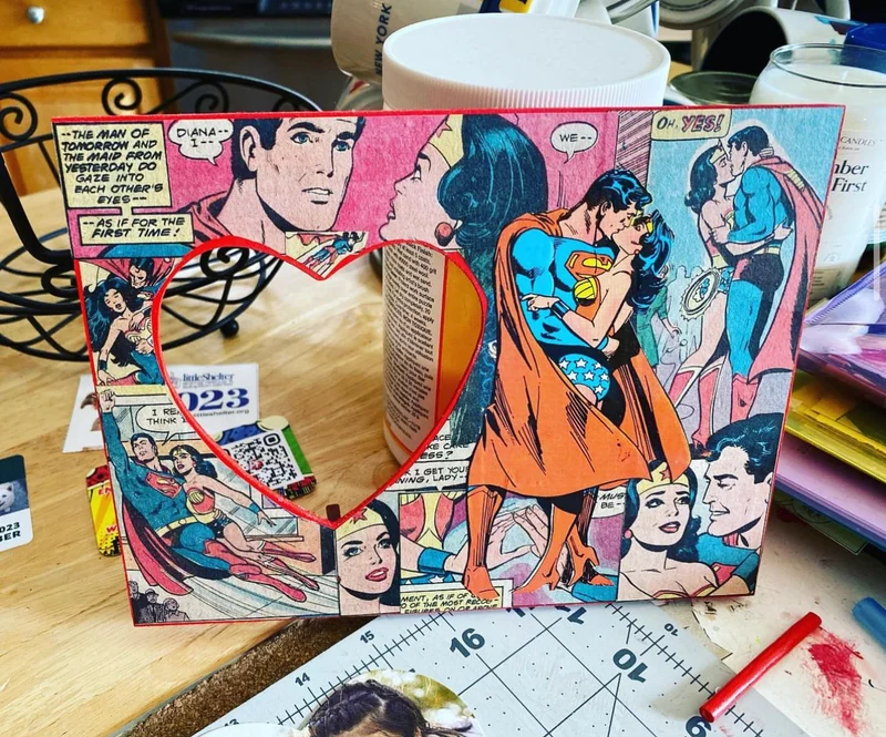 A handmade comic-panel photo frame with a heart-shaped opening and Superman and Wonder Woman artwork.